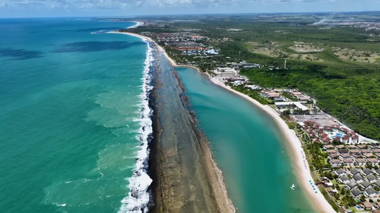 Foto aérea de uma faixa costeira longa e estreita, separando o oceano agitado à esquerda de águas calmas e esverdeadas à direita, com vegetação e construções ao fundo.