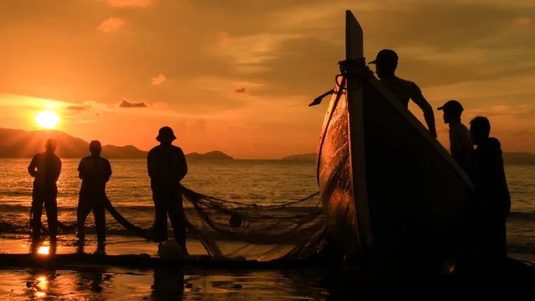 Silhueta de seis pescadores em um barco de pesca tradicional durante o pôr do sol, com o céu em tons alaranjados e reflexos dourados na água.